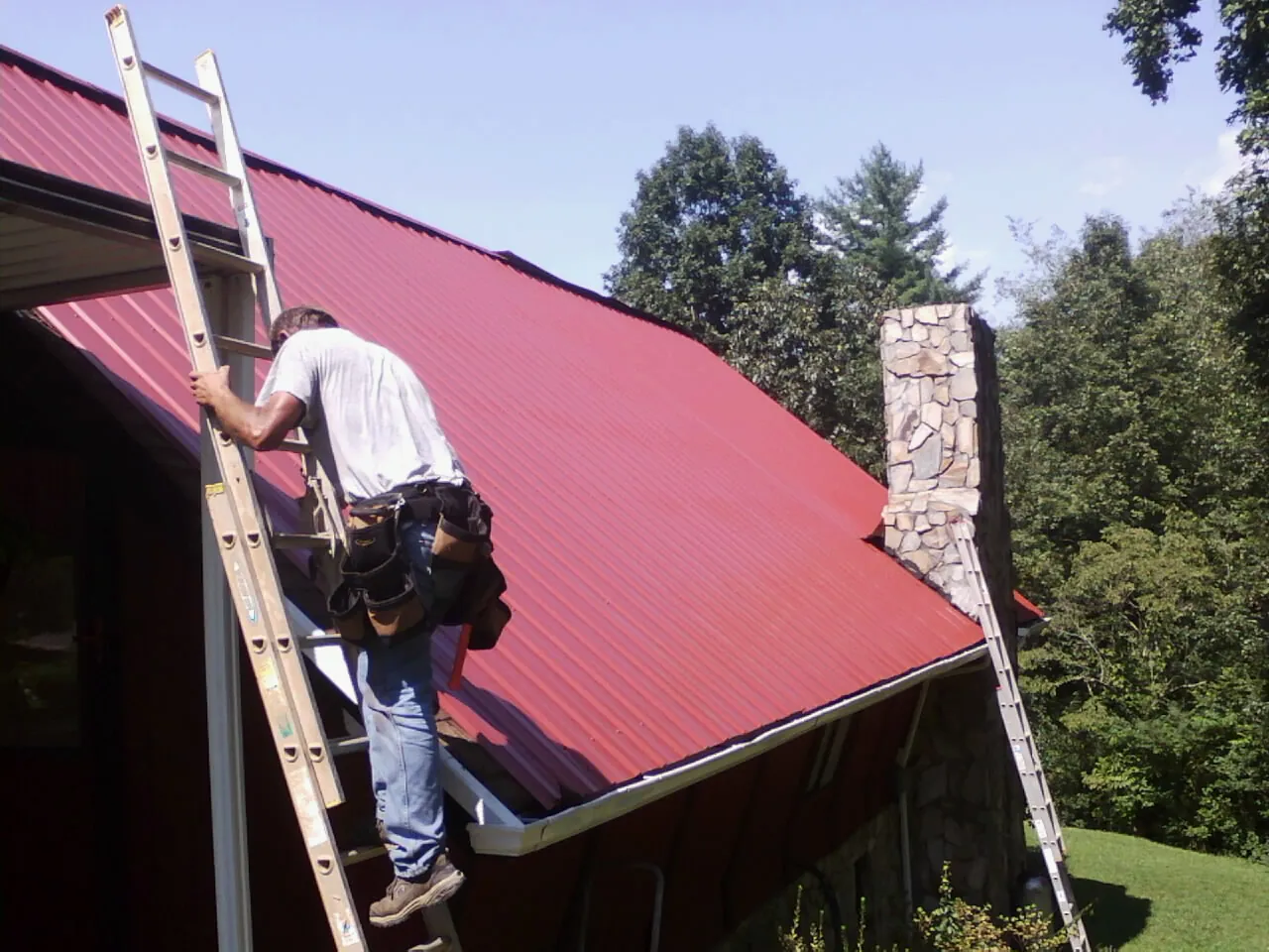 Roofer on a ladder beside a red metal roof and stone chimney — Soffit & Fascia Repair by Roofing Brothers in Crittenden